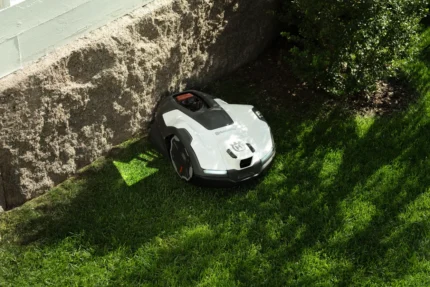 Robotic lawn mower operating on grass near a stone wall and bushes.