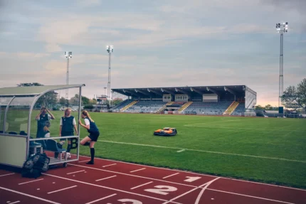 Ein Mähroboter, der auf einem Sportplatz in der Nähe einer Laufbahn arbeitet, mit einem Stadion im Hintergrund.