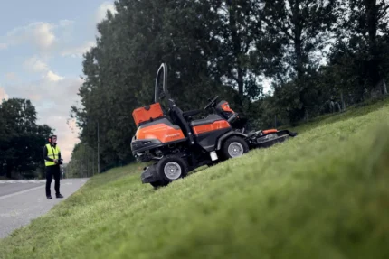 Eine Person, die einen Aufsitzmäher beobachtet, der an einem Hang neben einer Straße Gras mäht.