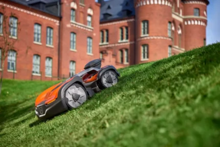 Mähroboter auf einer grasbewachsenen Steigung mit einem historischen Gebäude im Hintergrund.