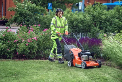 Eine Person in Arbeitskleidung, die einen Rasenmäher verwendet, um Gras in einer Gartenumgebung zu schneiden.