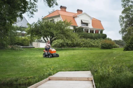 Eine Person, die mit einem Rider-Rasenmäher in einem Garten mit einem großen Haus im Hintergrund einen Rasen mäht.