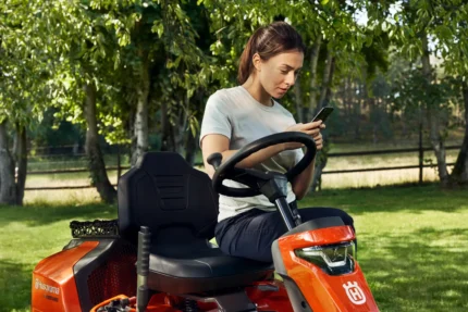 Eine Person, die auf einem Husqvarna Aufsitzmäher in einem Garten sitzt und ein Smartphone in der Hand hält.