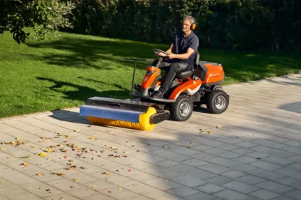 Eine Person, die eine Aufsitzkehrmaschine verwendet, um Blätter von einer gepflasterten Fläche neben einem Rasen zu entfernen.