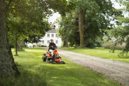 Eine Person, die mit einem Aufsitzmäher den Rasen in einer Gartenumgebung mit Bäumen und einem Haus im Hintergrund mäht.